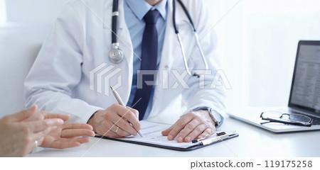 A doctor and a patient. The physician, wearing a white medical coat over a blue shirt and tie, is filling out a medical record form during a consultation in the clinic. Medical service A doctor and a patient. The physician, wearing a white medical coat over a blue shirt and tie, is filling out a medical record form during a consultation in the clinic. Medical service 119175258