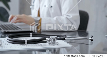 Stethoscope and clipboard with medication history records are on a glass desk behind a physician using a laptop computer. Medicine concept Stethoscope and clipboard with medication history records are on a glass desk behind a physician using a laptop computer. Medicine concept 119175290