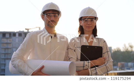 Man and woman architects, wearing safety helmets, are standing near each other and looking at the camera at a construction site during sunrise or sunset. Architecture and engineering concept 119175294