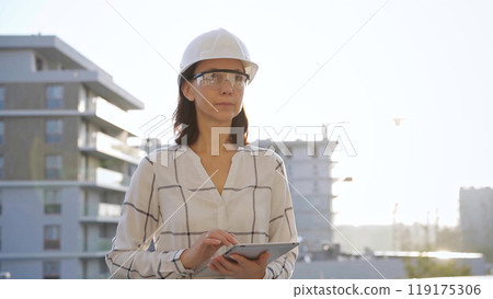 Woman construction engineer wearing white checked blouse and hard hat is using tablet computer while inspecting a building site at sunset, front view. Architecture and engineering concepts Woman construction engineer wearing white checked blouse and hard hat is using tablet computer while inspecting a building site at sunset, front view. Architecture and engineering concepts 119175306