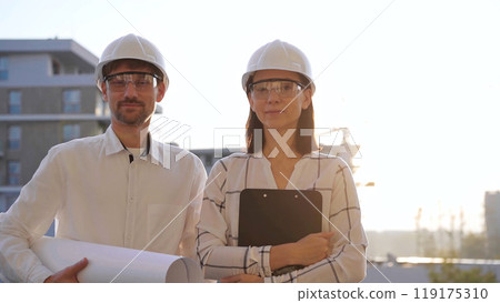 Man and woman architects, wearing safety helmets, are standing near each other and looking at the camera at a construction site during sunrise or sunset. Architecture and engineering concept Man and woman architects, wearing safety helmets, are standing near each other and looking at the camera at a construction site during sunrise or sunset. Architecture and engineering concept 119175310