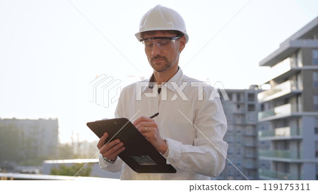 Man construction engineer wearing white shirt and hard hat is making notes on a clipboard while inspecting a building site at sunset, front view. Architecture and engineering concepts 119175311