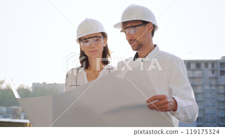 Two architects, wearing safety helmets and glasses, are examining blueprints at a construction site during sunrise or sunset, front view. Architecture and engineering concepts Two architects, wearing safety helmets and glasses, are examining blueprints at a construction site during sunrise or sunset, front view. Architecture and engineering concepts 119175324