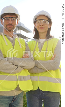 Man and woman construction engineers wearing hard hats and safety vests are standing with arms crossed, smiling at the camera with a construction site and sunrise at the background. Vertical portrait Man and woman construction engineers wearing hard hats and safety vests are standing with arms crossed, smiling at the camera with a construction site and sunrise at the background. Vertical portrait 119175357
