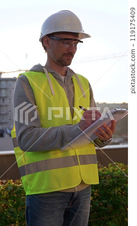 Man engineer with white helmet and safety vest is taking notes on a clipboard while inspecting a construction site at sunrise in early morning, vertical front view. Architecture and engineering Man engineer with white helmet and safety vest is taking notes on a clipboard while inspecting a construction site at sunrise in early morning, vertical front view. Architecture and engineering 119175409