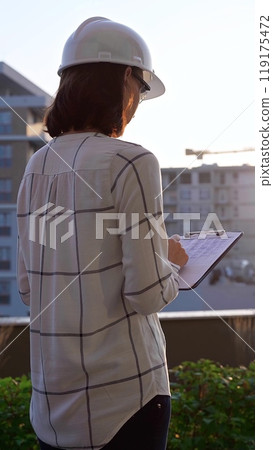Woman construction engineer wearing white checked blouse and hard hat is making notes on a clipboard while inspecting a building site at sunset, back and vertical view. Architecture concept Woman construction engineer wearing white checked blouse and hard hat is making notes on a clipboard while inspecting a building site at sunset, back and vertical view. Architecture concept 119175472