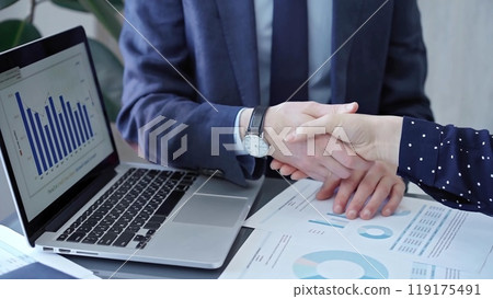 Two business people professionals shaking hands in office setting, sealing a deal with laptop and financial graphs visible. Business handshake over laptop and reports 119175491