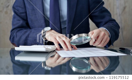 Auditor man analyzing financial documents with magnifying glass and calculator at his glass office desk in formal blue jacket. Business people concept 119175512