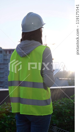 Woman construction engineer with a white hard hat and safety vest is using digital tablet while inspecting a construction site in the early morning at sunrise, back view 119175541