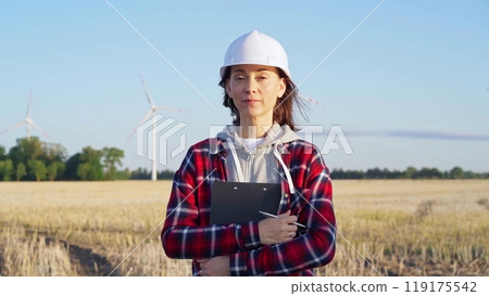 Woman engineer wearing white protective helmet is standing straight in a field with wind turbines, as the sun sets. Concept of clean energy and engineering audit Woman engineer wearing white protective helmet is standing straight in a field with wind turbines, as the sun sets. Concept of clean energy and engineering audit 119175542