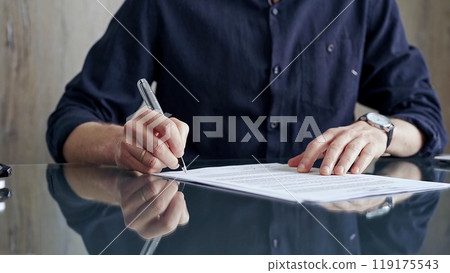 Businessman in dark blue t-shirt is signing contract at desk. Close-up of a male executive signing a legal document on a glass desk. Business people concept Businessman in dark blue t-shirt is signing contract at desk. Close-up of a male executive signing a legal document on a glass desk. Business people concept 119175543