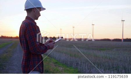 Man engineer wearing a white protective helmet is taking notes with a clipboard in a field with wind turbines, as the sun sets. Clean energy and engineering concept 119175639