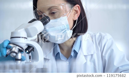 Female scientist in lab goggles, protective mask and gloves is using microscope in laboratory. Science concept Female scientist in lab goggles, protective mask and gloves is using microscope in laboratory. Science concept 119175682