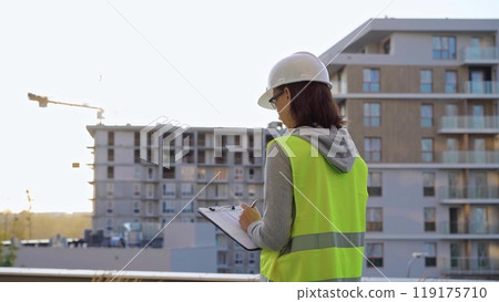 Woman engineer with a hard hat and safety vest is writing on a clipboard while inspecting a construction site at sunset Woman engineer with a hard hat and safety vest is writing on a clipboard while inspecting a construction site at sunset 119175710