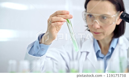 Woman scientist with protective glasses examining lab tube with a green fluid. Microbiology and science concept 119175738
