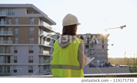 Female construction engineer with a hard hat and safety vest is writing on a clipboard while inspecting a construction site in the evening at sunset Female construction engineer with a hard hat and safety vest is writing on a clipboard while inspecting a construction site in the evening at sunset 119175739