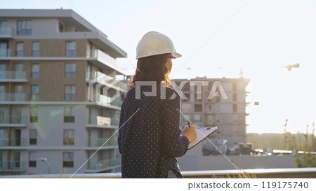 Woman architect wearing a hard hat is taking notes with a tablet computer while inspecting a building construction site at sunset 119175740