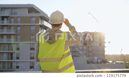 Male engineer with white hard hat and safety vest is using a tablet computer while inspecting a construction site at sunset 119175779