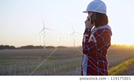 Woman engineer talking on the phone in a field with wind turbines, as the sun sets. Concept of clean energy and engineering 119175801