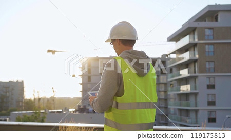Man constructive engineer with white hard hat and safety vest is using a tablet computer while inspecting a construction site at sunset 119175813
