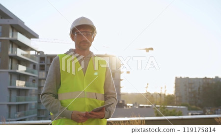 Construction site engineer holding tablet and wearing hard hat and safety vest with a sunrise at the background. Portrait video Construction site engineer holding tablet and wearing hard hat and safety vest with a sunrise at the background. Portrait video 119175814