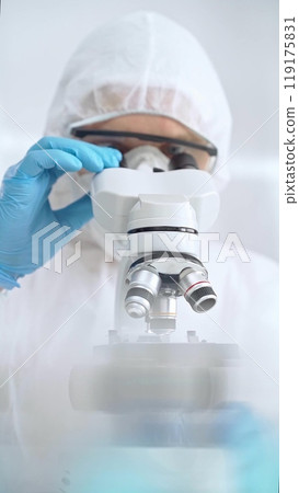 Scientist in ppe analyzing samples with microscope. Close-up of a focused researcher in a lab coat and protective gear using a microscope in a laboratory setting 119175831