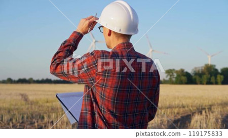 Man engineer at work is wearing a white protective helmet and taking notes with a clipboard in a field with wind turbines, as the sun sets. Clean energy concept Man engineer at work is wearing a white protective helmet and taking notes with a clipboard in a field with wind turbines, as the sun sets. Clean energy concept 119175833