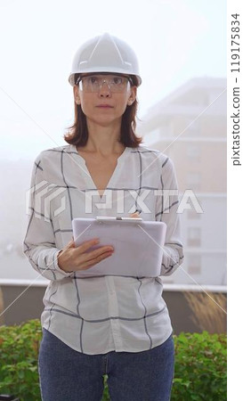 Female construction engineer wearing white checked blouse and safety helmet is making notes on a clipboard while inspecting a building site in foggy weather, front vertical view. Architecture concept 119175834