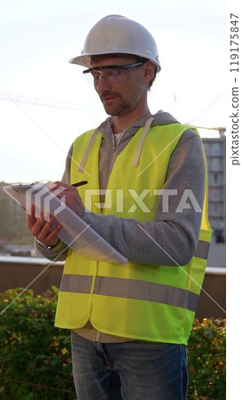 Man engineer with white hard hat and safety vest is taking notes on a clipboard while inspecting a construction site at sunrise, vertical front view. Architecture and engineering Man engineer with white hard hat and safety vest is taking notes on a clipboard while inspecting a construction site at sunrise, vertical front view. Architecture and engineering 119175847