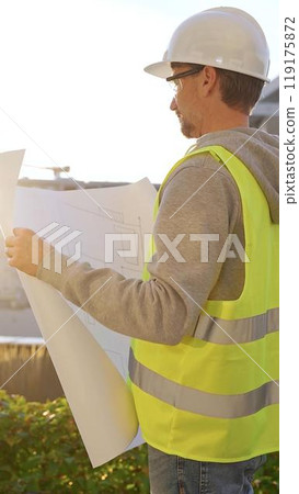 Male architect wearing safety hard hat and vest holding blueprint and looking to construction site at sunrise, back vertical view. Architecture and engineering Male architect wearing safety hard hat and vest holding blueprint and looking to construction site at sunrise, back vertical view. Architecture and engineering 119175872