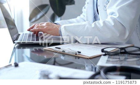 Doctor man with blue shirt and medical coat is using laptop computer in clinic office while sitting at the glass desk. Medicine concept Doctor man with blue shirt and medical coat is using laptop computer in clinic office while sitting at the glass desk. Medicine concept 119175873