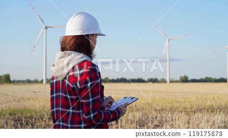 Female engineer wearing a white protective helmet is taking notes with a clipboard in a field with wind turbines, as the sun sets. Clean energy concept Female engineer wearing a white protective helmet is taking notes with a clipboard in a field with wind turbines, as the sun sets. Clean energy concept 119175878