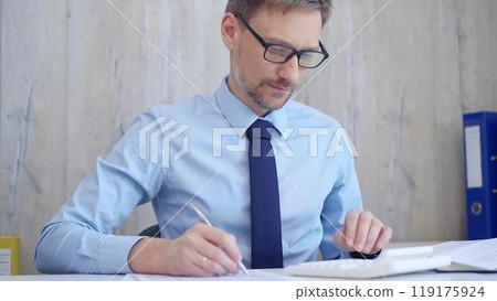 Male accountant with blue t-shirt concentrating while calculating costs and taking notes at his desk. Taxes, audit in business 119175924