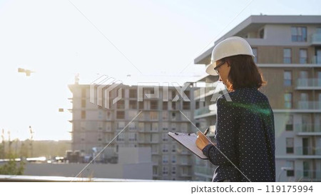 Woman architect wearing a hard hat is taking notes with a tablet computer while inspecting a building construction site at sunset Woman architect wearing a hard hat is taking notes with a tablet computer while inspecting a building construction site at sunset 119175994