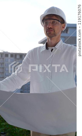 Construction site manager, wearing helmets and safety glasses, is holding blueprints and inspecting a building site during sunrise or sunset, front view vertical portrait. Architecture and engineering Construction site manager, wearing helmets and safety glasses, is holding blueprints and inspecting a building site during sunrise or sunset, front view vertical portrait. Architecture and engineering 119176076