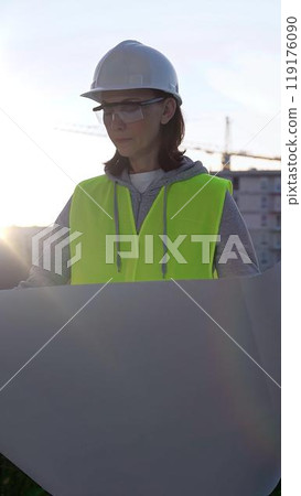 Female architect or engineer wearing safety hard hat and vest holding blueprint while inspecting a construction site at sunrise in early morning, front vertical view. Architecture concept 119176090