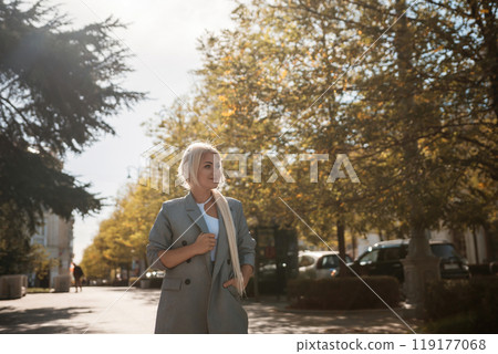 A woman wearing a suit and a scarf walks down a street. The street is lined with trees and parked cars. 119177068