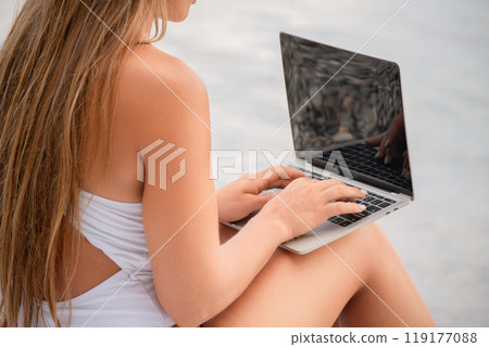 A woman is sitting on a beach with a laptop in her lap. She is wearing headphones and she is listening to music. A woman is sitting on a beach with a laptop in her lap. She is wearing headphones and she is listening to music. 119177088
