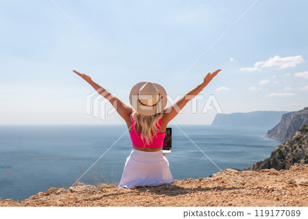 A woman is sitting on a rock by the ocean with a laptop open in front of her. She is wearing a pink top and a straw hat. Concept of relaxation and leisure. A woman is sitting on a rock by the ocean with a laptop open in front of her. She is wearing a pink top and a straw hat. Concept of relaxation and leisure. 119177089