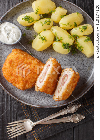 Schnitzel cordon bleu is a dish of meat with cheese and ham filling, then breaded and pan-fried served with new potatoes and cream sauce closeup on the plate. Vertical top view 119177120