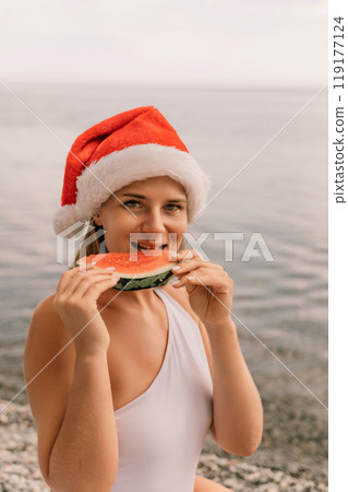 A woman is eating a watermelon while wearing a red santa hat. Scene is lighthearted and festive, as the woman is enjoying a refreshing snack on a sunny day. A woman is eating a watermelon while wearing a red santa hat. Scene is lighthearted and festive, as the woman is enjoying a refreshing snack on a sunny day. 119177124