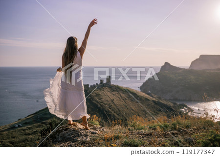 A woman in a white dress stands on a rocky hill overlooking the ocean. She is smiling and she is happy. 119177347