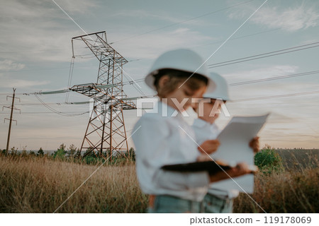 Little kids playing as engineers or managers with safety helmets, using tablet. Electrical industry profession, serious smart children on transmission power lines background. Little kids playing as engineers or managers with safety helmets, using tablet. Electrical industry profession, serious smart children on transmission power lines background. 119178069