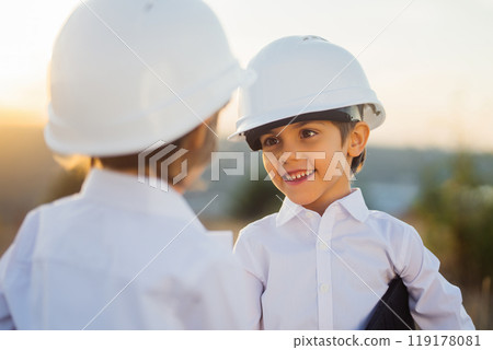 Little boys acting role of engineers or managers with safety helmet. Occupation in electrical industry, smiling smart children on electric voltage towers background Little boys acting role of engineers or managers with safety helmet. Occupation in electrical industry, smiling smart children on electric voltage towers background 119178081
