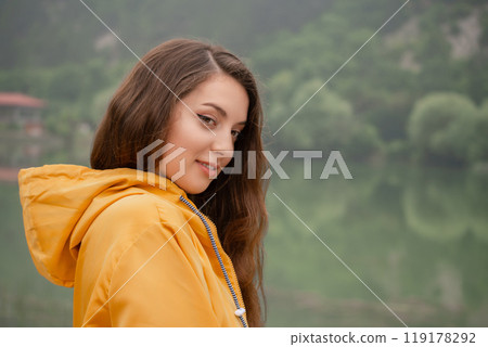 A woman in a yellow jacket stands in front of a lake A woman in a yellow jacket stands in front of a lake 119178292