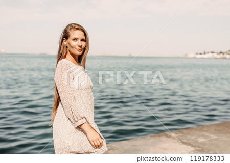 A woman is walking on the beach wearing a dress. The water is calm and the sky is clear. 119178333