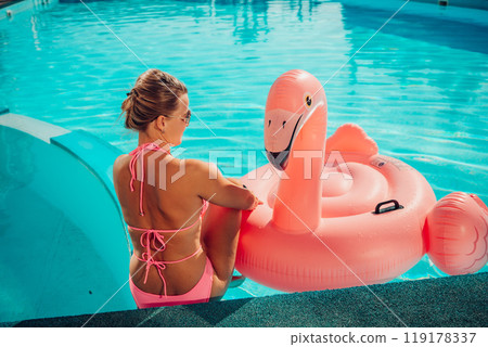A woman in a pink bikini is sitting on a pink inflatable flamingo in a pool. The scene is playful and fun, with the woman enjoying her time in the water. 119178337