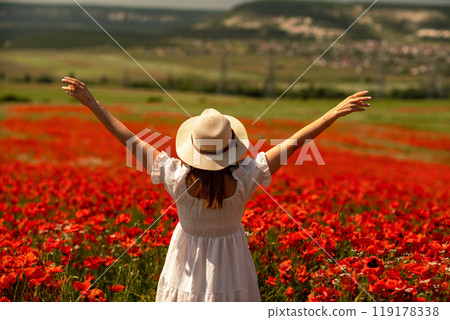 Field poppies woman. Happy woman in a white dress and hat stand through a blooming field of poppy raised her hands up. Field of blooming poppies. Field poppies woman. Happy woman in a white dress and hat stand through a blooming field of poppy raised her hands up. Field of blooming poppies. 119178338