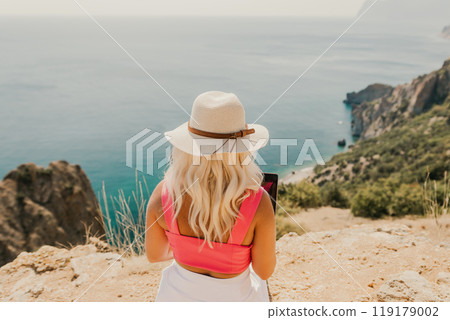 A woman is sitting on a rocky hillside overlooking the ocean. She is wearing a pink shirt and a white hat. A woman is sitting on a rocky hillside overlooking the ocean. She is wearing a pink shirt and a white hat. 119179002