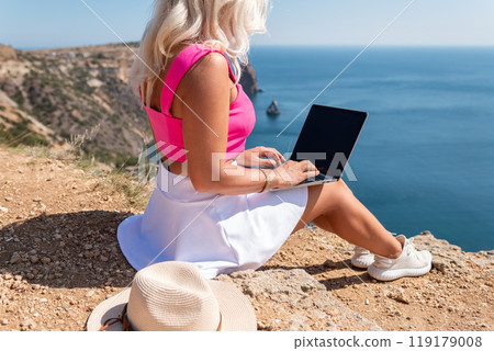 A woman is sitting on a rock by the water with a laptop open in front of her. She is typing on the laptop and she is focused on her work. Concept of productivity and concentration. 119179008